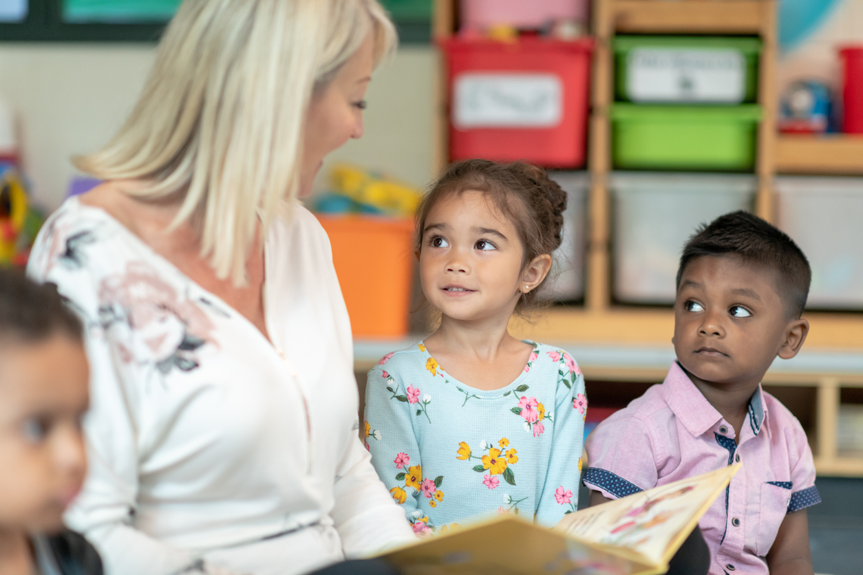 A preschool teacher is sitting on the floor with a group of students in her classroom. The female educator is reading to the preschool kids. The multi-ethnic group of children is huddled around the teacher listening and smiling.