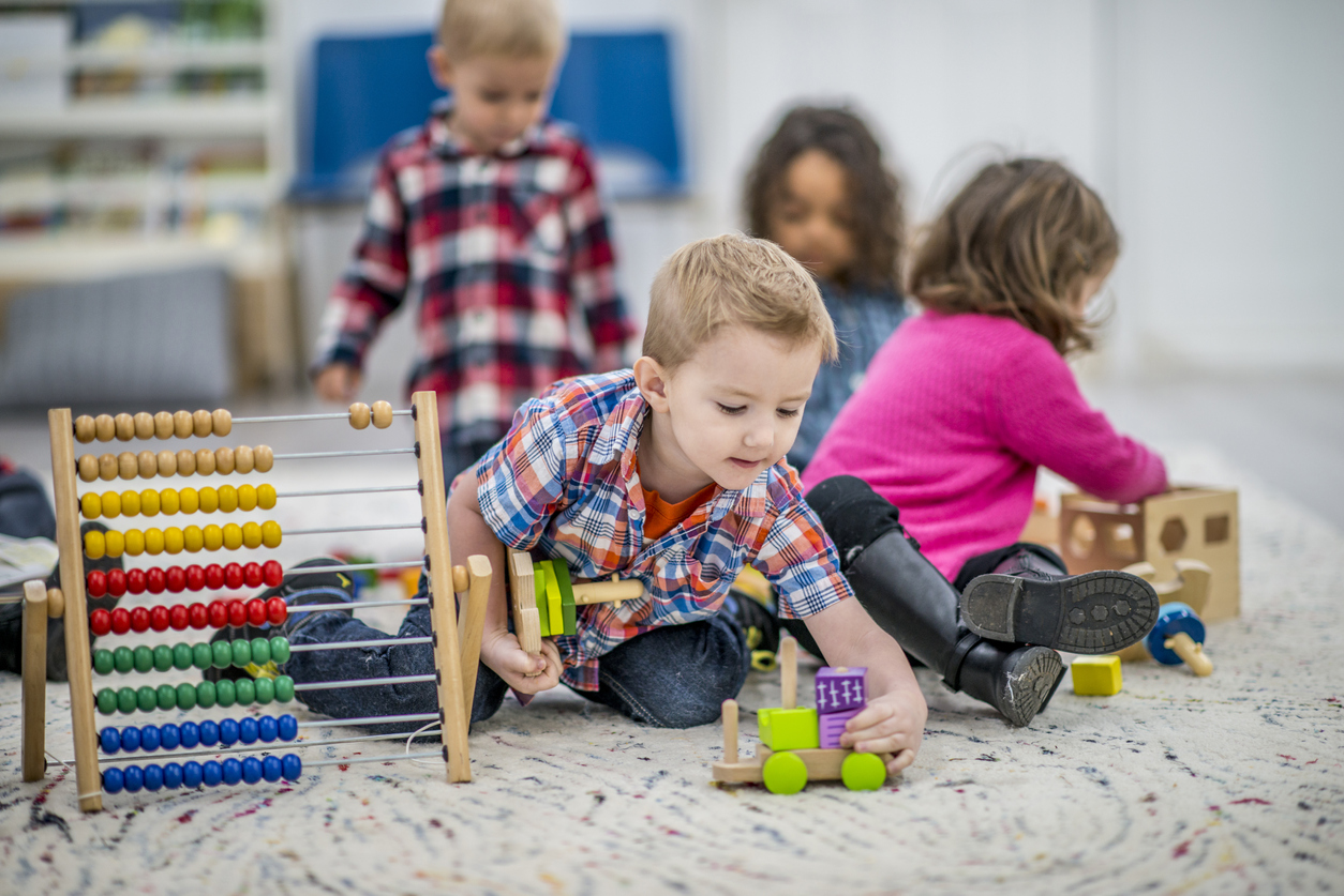 A multi-ethnic group of young children are indoors in a preschool. They are wearing casual clothing. In the foreground, a Caucasian boy is playing with a toy car and abacus.